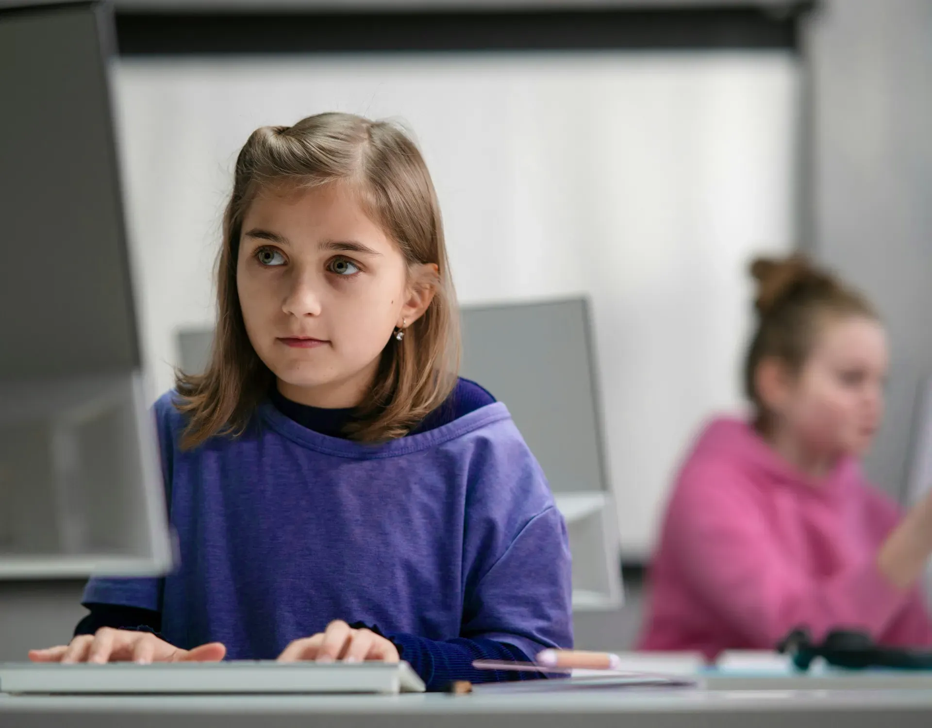 A young girl sits attentively at a computer in a classroom, with a classmate working quietly in the background.