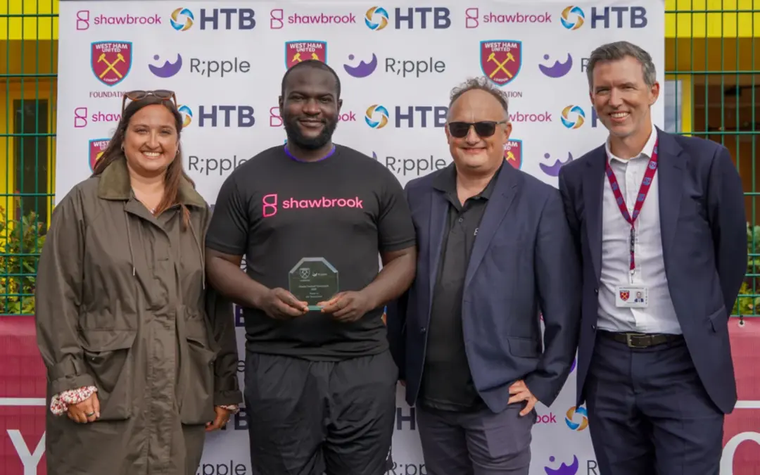 Cuatro personas en el torneo de Ripple y West Ham United Foundation, una de ellas sosteniendo un premio de cristal y vistiendo una camiseta de Shawbrook.