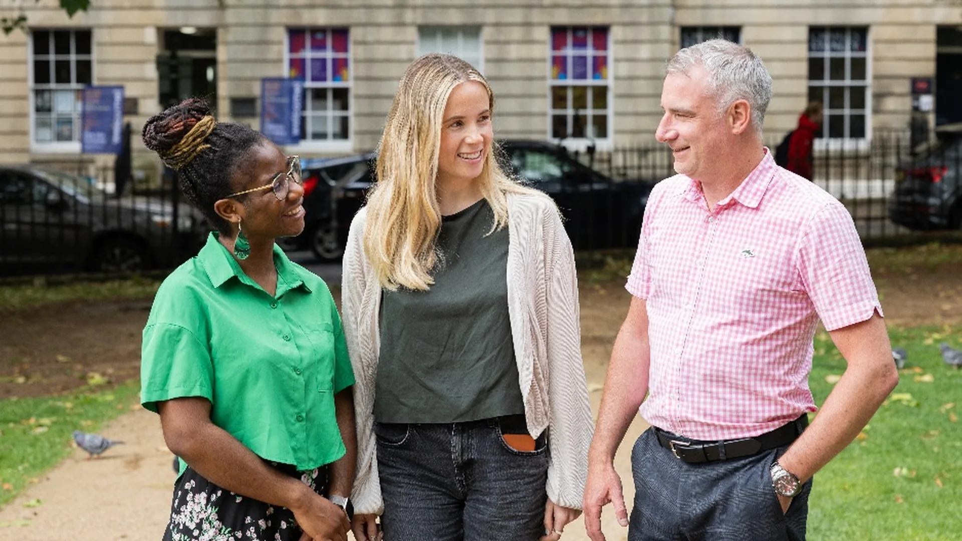 Three people smiling and talking on a park path in front of a Georgian stone building in Bath
