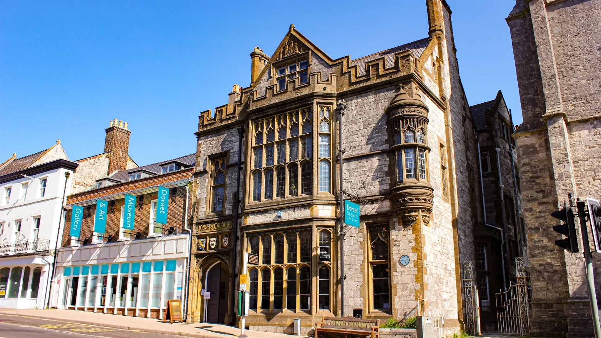 Gothic-style stone frontage of Dorset Museum & Art Gallery on a sunny day, with teal banners on the neighbouring building